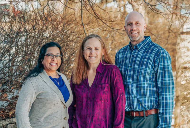 The image shows three individuals posing together outdoors during daylight they appear to be at an event, possibly a formal occasion given their attire, with one person wearing a tie.