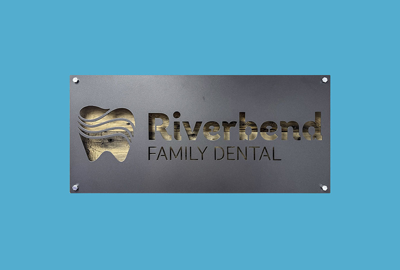 The image features a sign with text that reads RIVERBEND FAMILY DENTAL on a blue background, set against a backdrop of a building facade.