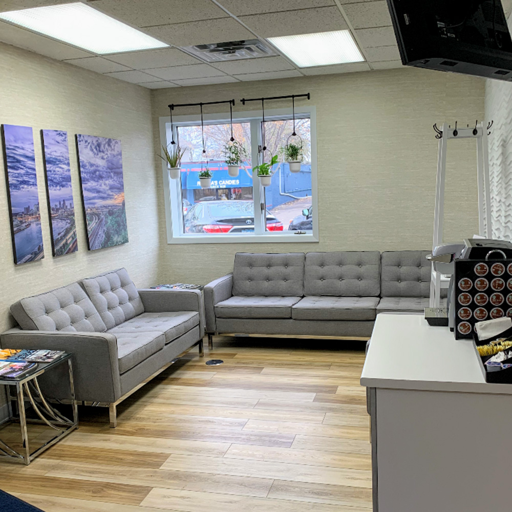 The image shows an interior space with a waiting area featuring a couch, a television, and a reception desk. There are framed pictures on the wall above the couch, and a large window with a view of a cityscape can be seen through the window to the right. The room has a modern design with neutral tones and is well-lit.