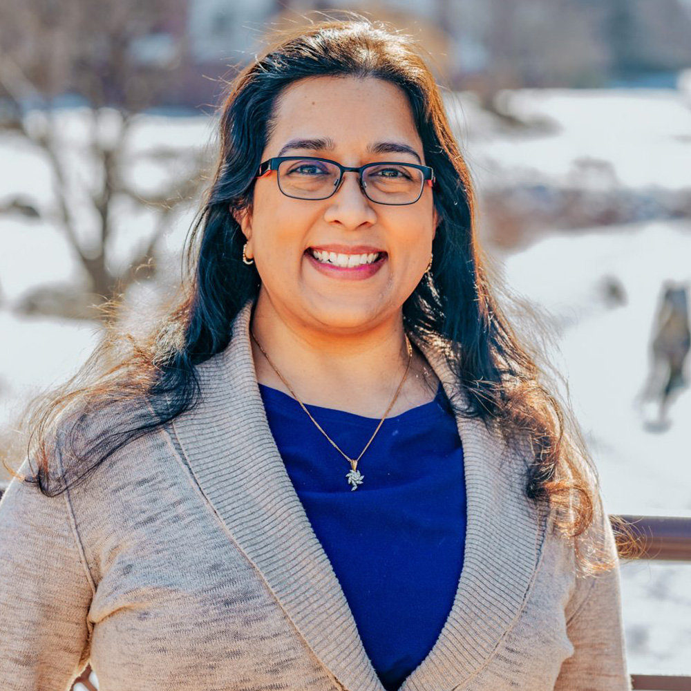 A woman with glasses smiling at the camera, wearing a blue shirt, standing outdoors under a clear sky, dressed in a light-colored jacket over a dark top, with a snowy landscape in the background.