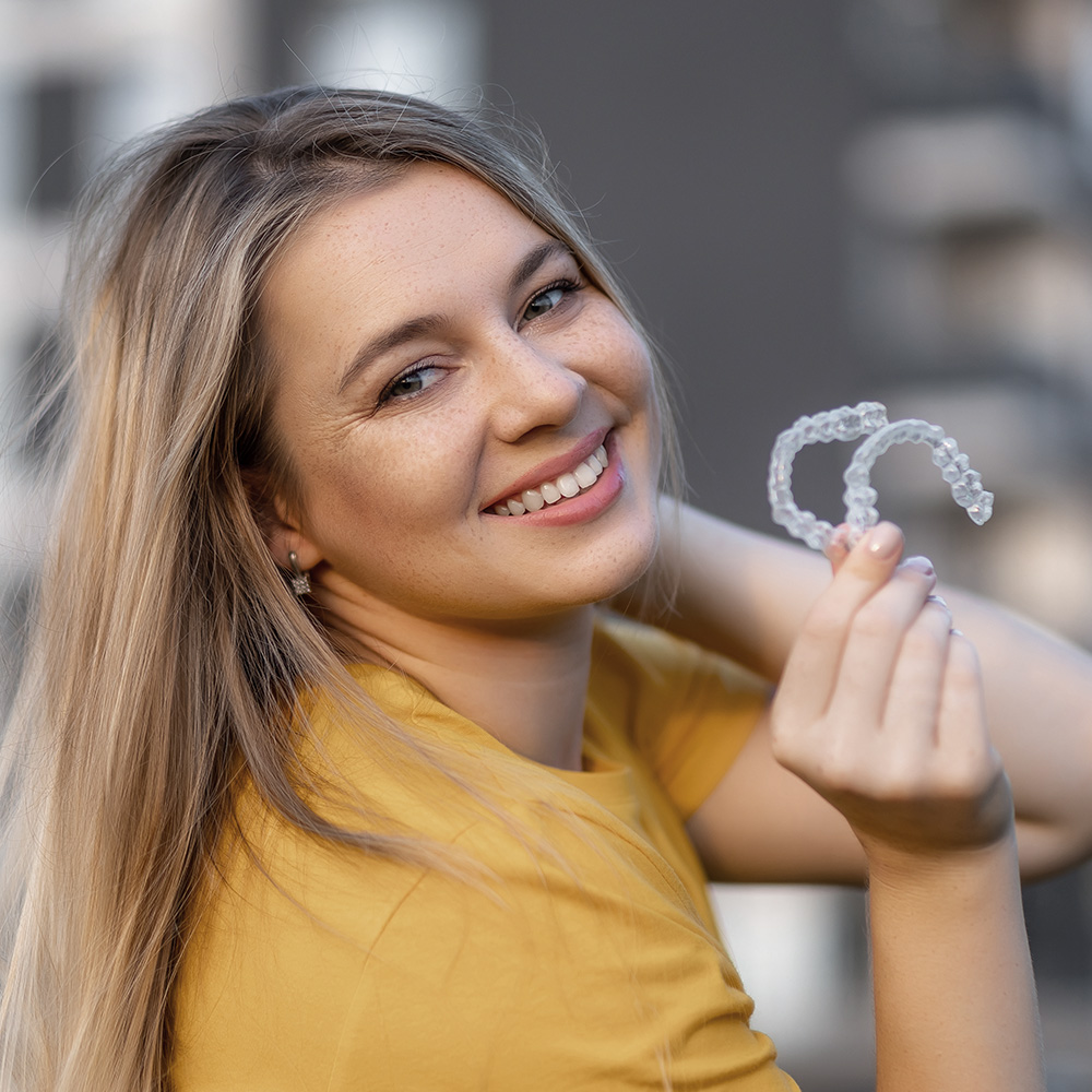 The image shows a smiling woman with long blonde hair holding up a clear plastic object shaped like a toothbrush, possibly a dental appliance, against her face. She s outdoors wearing a yellow top and has one hand on her head.